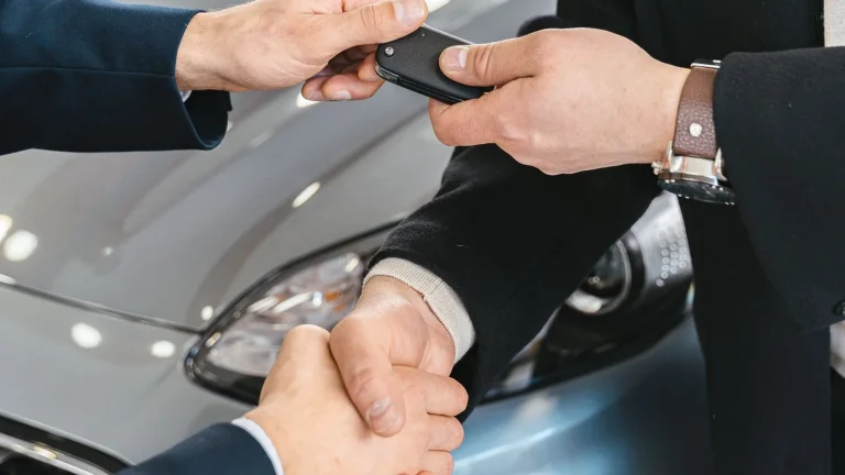 Two people shaking hands in a car dealership while one hands over a car key